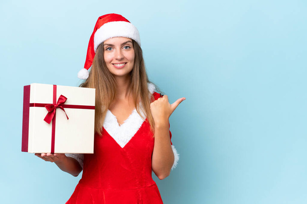 Anne Noel gibi giyinmiş, mavi arka planda izole edilmiş beyaz bir kadın bir ürünü sunmak için kenarı işaret ediyor. - Fotoğraf, Görsel