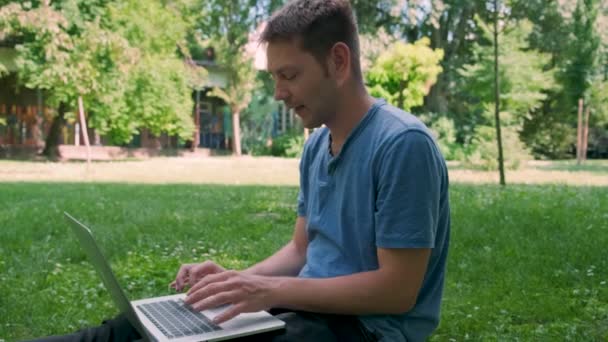 Mies freelancer sininen t-paita istua vihreä ruoho kaupungin puistossa, työskennellä kannettavan tietokoneen ja juo kahvia paperimuki. Miesoppilas koneella ulkona. Etätyö, verkko-opetuksen käsite. - Materiaali, video