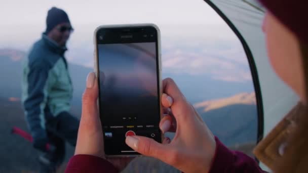 Kobieta robi zdjęcie turystce w pięknej scenerii górskiej przy użyciu telefonu. Afroamerykanin z siekierą w ręku pozuje na wzgórzu. Backpackers zatrzymał się odpocząć podczas wakacji przygody. Zamknij się.. - Materiał filmowy, wideo