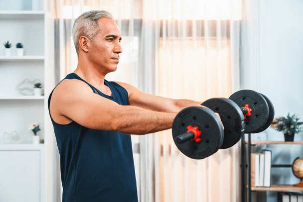 Atletische en sportieve senior man die zich bezighouden met lichaamsbeweging routine met tillen halter thuis als concept van gezonde fit lichaam met lichaamsgewicht levensstijl na pensionering. Klok - Foto, afbeelding