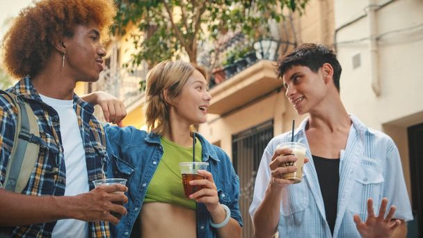 Группа счастливых друзей, два парня и девушка, гуляющие с выпивкой на вынос вдоль городской улицы, болтая и смеясь - Фото, изображение