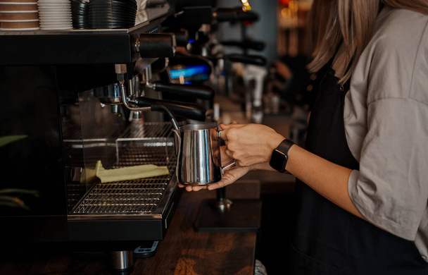 Gros plan d'une main de baristas versant du lait moussé dans une tasse à café en papier, avec une machine à expresso floue en arrière-plan - Photo, image