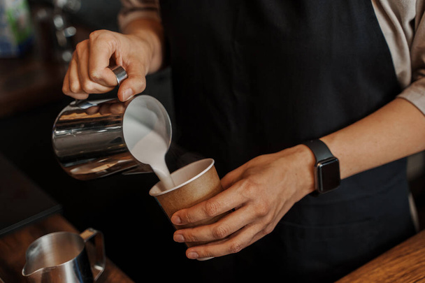Primo piano delle mani di un barista che versano il latte schiumato in una tazza di caffè di carta, con una macchina da caffè espresso sfocata sullo sfondo - Foto, immagini