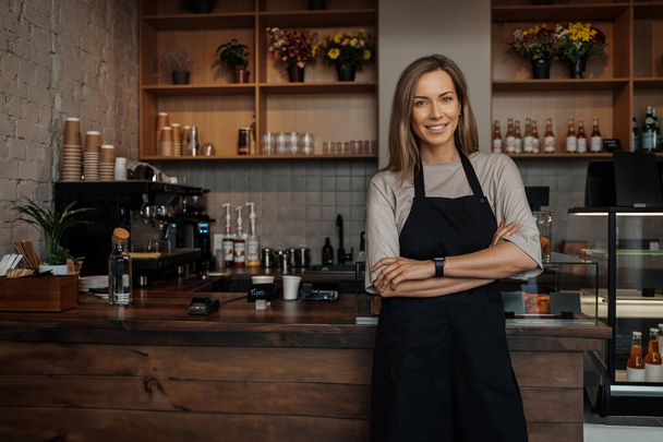 Smiling woman in apron standing confidently in a cozy cafe environment, with shelves of flowers in the background - Photo, Image