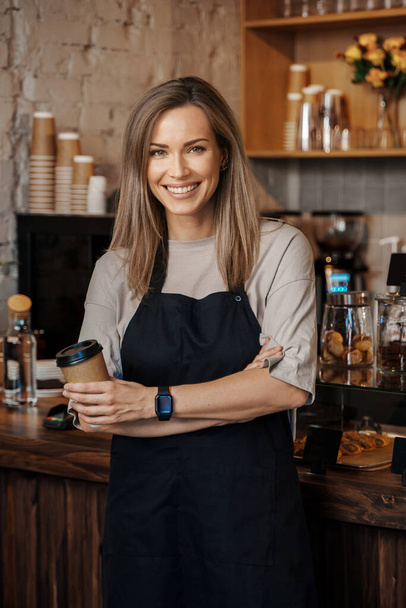 Ritratto di una barista in bilico con in mano una tazzina da asporto, in piedi nel suo moderno caffè - Foto, immagini