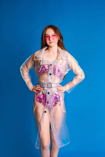 teenager girl wearing in swimsuit and transparent raincoat on blue background, ready for tropical rainy showers - Photo, Image