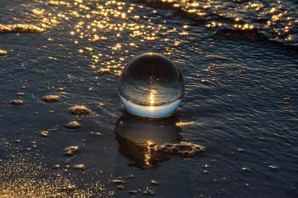 Eine Glaskugel liegt in den Wellen am Sandstrand, das Meer und die untergehende Sonne spiegeln sich in der Kugel - Foto, Bild