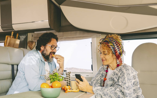Couple of adult tourist enjoy leisure time together inside a modern camper van. Renting motor home for summer travel holiday vacation. Van lifestyle for young people. Woman using phone and man eating - Foto, imagen