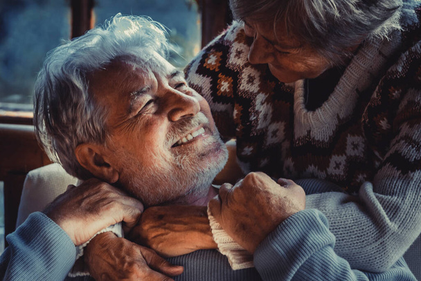 Portret van een stel oudere mensen die thuis verliefd zijn in het donker. Oude man glimlacht naar haar volwassen vrouw die knuffelt en geniet van een relatie. Winter thuis achtergrond. Ouderen leven - Foto, afbeelding