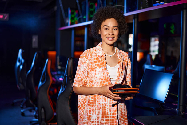 happy african american woman wearing casual clothing holding headphones in cybersport club - Photo, Image