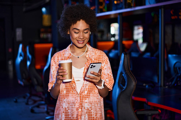 stylowa Afroamerykanka trzyma filiżankę kawy i jej telefon i uśmiecha się w pomieszczeniach - Zdjęcie, obraz
