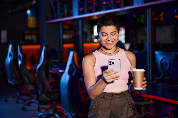 stylish woman, clad in fashionable clothing, smiles as she holds her phone and a cup of coffee - Photo, Image