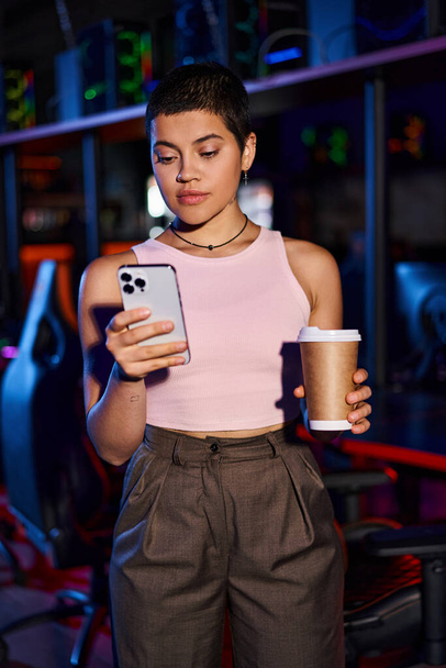 Μια νεαρή γυναίκα στέκεται σε εσωτερικούς χώρους, όπως αυτή κρατώντας ένα τηλέφωνο και καφέ χάρτινο κύπελλο στο Cybersport Club - Φωτογραφία, εικόνα