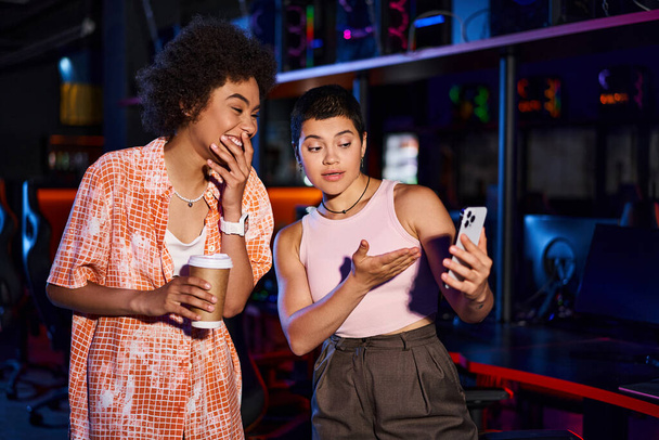 Kaksi tyylikästä rotujenvälinen naiset jakavat hetki iloa ja yhteyden kahvin yli, katsot puhelinta - Valokuva, kuva