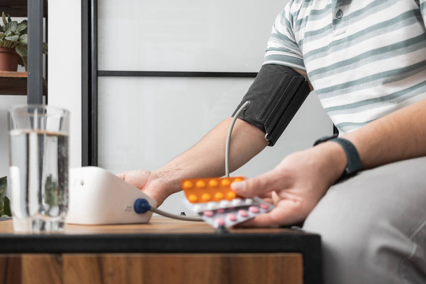 Неузнаваемый мужчина сидит дома на диване и измеряет артериальное давление на руке современным цифровым тонометром и держит пузырьки с таблетками, лекарствами . - Фото, изображение