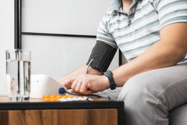 Неузнаваемый мужчина сидит дома на диване и измеряет артериальное давление на руке современным цифровым тонометром. - Фото, изображение
