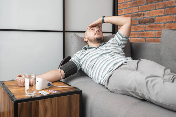 Un jeune homme malsain souffrant d'hypertension et de maux de tête, allongé sur le canapé à la maison, mesure la pression artérielle sur sa main avec un tonomètre numérique moderne. - Photo, image