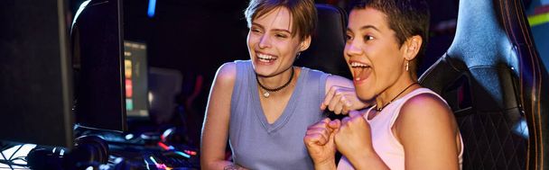 donne eccitate con tatuaggi che si concentrano su un gioco di cybersport nel club di computer, banner di giocatori femminili - Foto, immagini