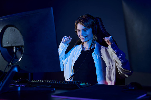 heureuse femme aux cheveux courts regardant l'ordinateur dans une pièce éclairée en bleu, concept de cybersport et de jeu - Photo, image