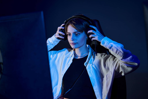 mujer enfocada con auriculares y mirando a la computadora en una habitación con luz azul, concepto de juego de ciberdeporte - Foto, imagen