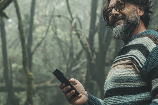 Ormanda tek başına duran adam gülümsüyor ve mobil uygulama bağlantısı kullanıyor. Veri teknolojisinde geziniyor. Dışarıdaki boş zamanlardaki insanlar. Turist doğanın tadını çıkarıyor ulusal park manzaralı bir yer - Fotoğraf, Görsel