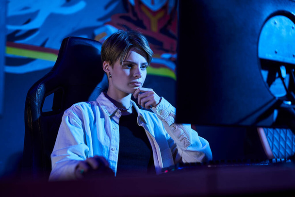 pensive woman with short hair looking at computer in a blue-lit room, cybersport game strategy - Photo, Image