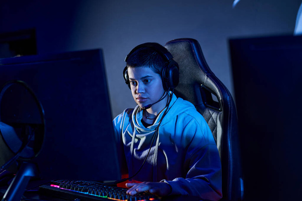 fokussierter Spieler mit kurzen Haaren, der in einem blau beleuchteten Raum auf den Computer schaut, Cybersport-Spieler im Kapuzenpulli - Foto, Bild