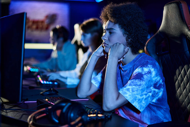 triste femme afro-américaine regardant le moniteur d'ordinateur et gonflant les joues dans la pièce éclairée bleue - Photo, image