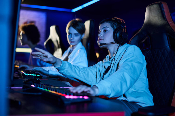 focus op gerichte vrouw met kort haar kijken naar computer monitor in de buurt van vrouwelijke gamers in de kamer - Foto, afbeelding