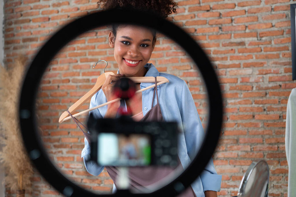 女性インフルエンサーがライブストリーミングビデオレビュー服を撮影 重要なソーシャルメディアやブログ. オンラインで録音セッション放送のためのアパレルスタジオ照明を持つ幸せな若い少女. - 写真・画像