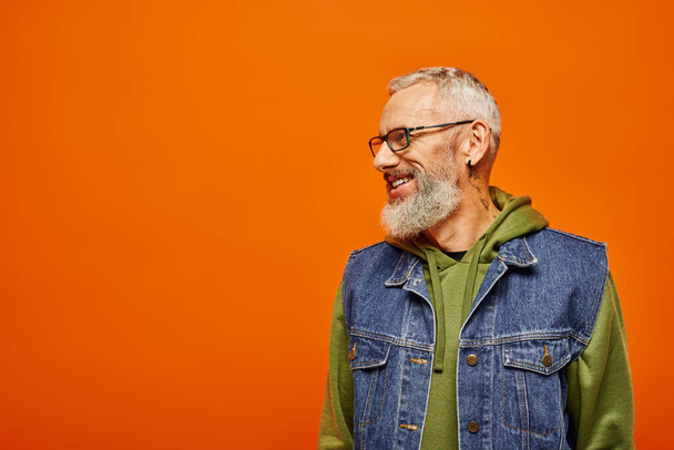 joyous handsome mature man in vibrant hoodie and vest with glasses smiling and looking away - Photo, Image