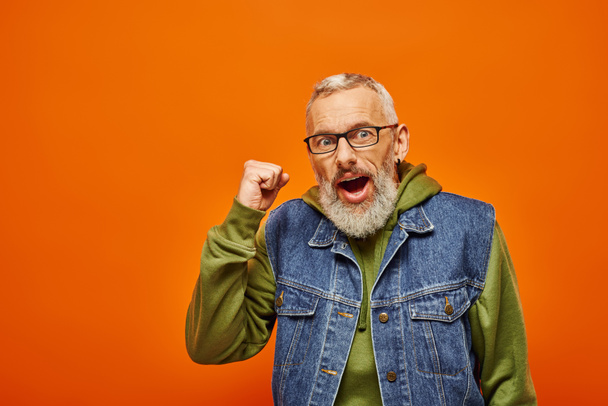 joyful mature funky man in green vibrant hoodie posing with fists near face on orange background - Photo, Image