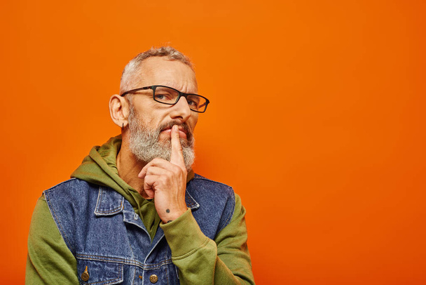 schöner, fokussierter älterer Mann in grünem Kapuzenpulli mit Brille und grauem Bart posiert auf orangefarbenem Hintergrund - Foto, Bild
