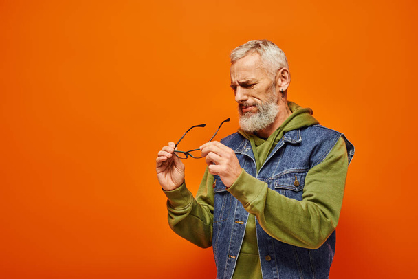 όμορφος συμπυκνωμένος ώριμος άντρας με ζωντανή εμφάνιση κρατώντας τα γυαλιά του στα χέρια σε πορτοκαλί φόντο - Φωτογραφία, εικόνα
