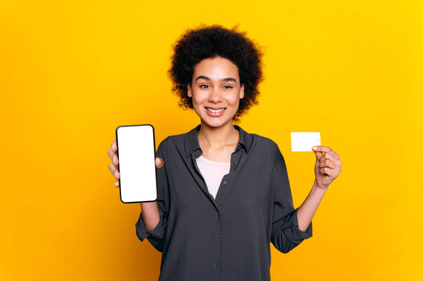 Positiv selbstbewusste hübsche afrikanisch-amerikanische Lockenkopf-Frau, stilvoll gekleidet, Bankberaterin, hält Handy und Kreditkarte in den Händen, blickt in die Kamera, lächelt, steht auf gelbem Hintergrund - Foto, Bild