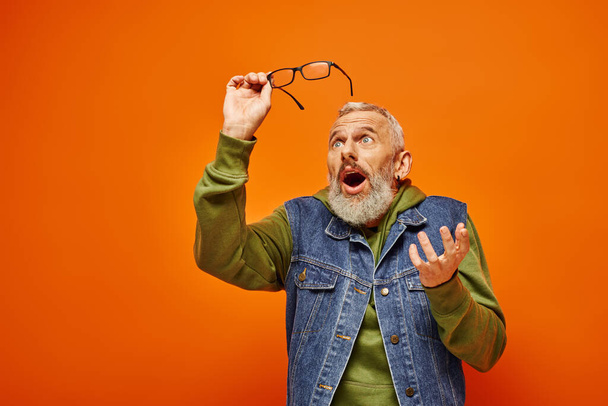 hombre maduro sorprendido de buen aspecto en traje vívido sosteniendo gafas y posando sobre fondo naranja - Foto, imagen
