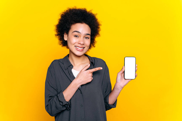 Ładna afrykańska lub brazylijska kobieta, patrzy w kamerę, trzyma w ręku smartfona z białym, pustym ekranem makietowym do prezentacji i wskazuje palcem na nią, stoi na odizolowanym żółtym tle, uśmiecha się - Zdjęcie, obraz