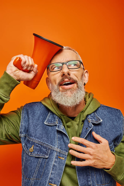 joyous surprised mature man in green hoodie and denim vest posing with megaphone on orange backdrop - Photo, Image