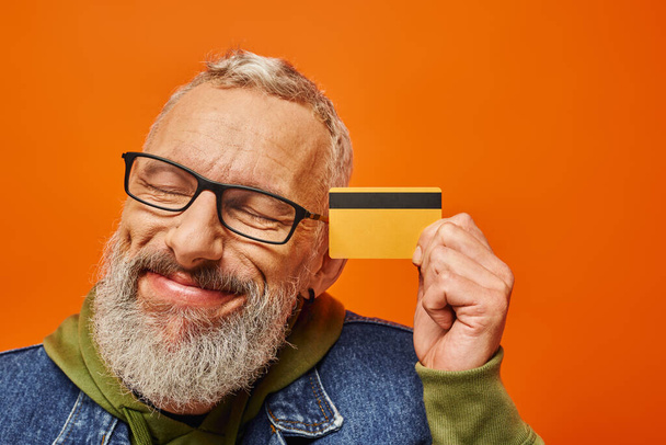 pohledný starší muž model v pulzujícím oblečení pózující se zavřenýma očima a držící kreditní kartu - Fotografie, Obrázek