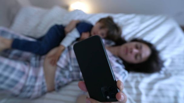 Padres e hijos usando pijamas, acostados en la cama y viendo contenido en el teléfono juntos - Foto, imagen