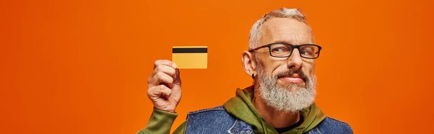 alegre bonito maduro homem em denim colete e capuz verde olhando para cartão de crédito na mão, banner - Foto, Imagem