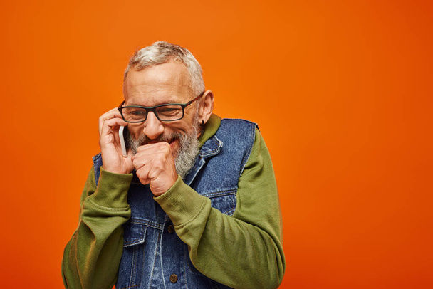 vrolijk knap volwassen man in trendy denim vest met baard en bril praten gelukkig per telefoon - Foto, afbeelding