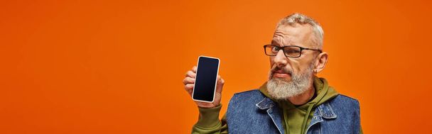confused good looking mature man in vibrant clothes holding phone and looking at camera, banner - Photo, Image