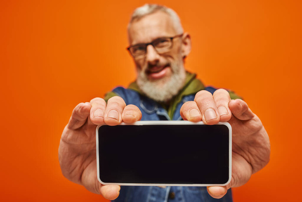 focus op smartphone in handen van vrolijke volwassen wazig man in levendige kleding op oranje achtergrond - Foto, afbeelding