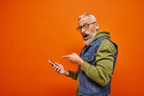 surprised handsome mature man in green hoodie pointing at smartphone and looking at camera - Photo, Image