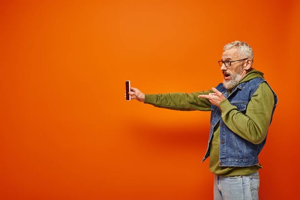 dobře vypadající starší mužský model v pulzujících šatech s vousy a brýlemi drží smartphone - Fotografie, Obrázek