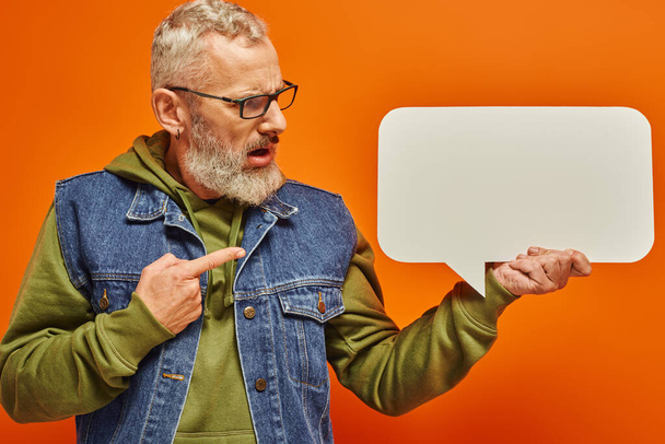 Verwirrter gutaussehender älterer Mann in Jeansweste blickt auf Sprechblase in der Hand vor orangefarbenem Hintergrund - Foto, Bild