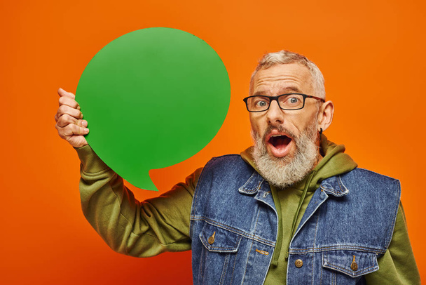shocked handsome mature male model in vibrant attire holding speech bubble and looking at camera - Photo, Image