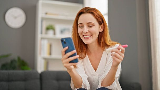 młoda ruda kobieta mając wideo połączenie trzymając ciąży test uśmiecha się w domu - Zdjęcie, obraz
