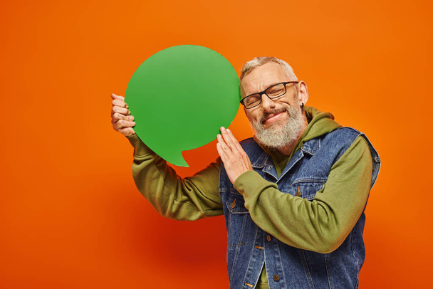 handsome joyous mature man with glasses and beard holding speech bubble and posing with closed eyes - Photo, Image